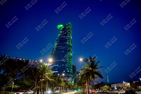 Evening view of a tall skyscraper illuminated with bright green lights in a city next to a road lined with palm trees. The sky is clear and dark blue, with car traffic moving along the road and streetlights illuminating the scene.