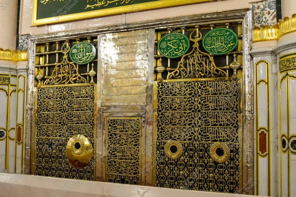 The image shows the exterior of the Prophet Muhammad\'s (PBUH) tomb grille in the Prophet\'s Mosque in Medina. The grille is made of iron adorned with gilded inscriptions that include Quranic verses and Islamic designs. At the top, there are green circular plaques with the names of significant Islamic figures.