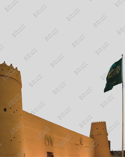 The image shows part of an ancient mudbrick fort with round towers featuring decorative holes. Also visible is the Saudi Arabian flag flying on a tall flagpole beside the fort. The sky is clear, providing a calm and simple background.