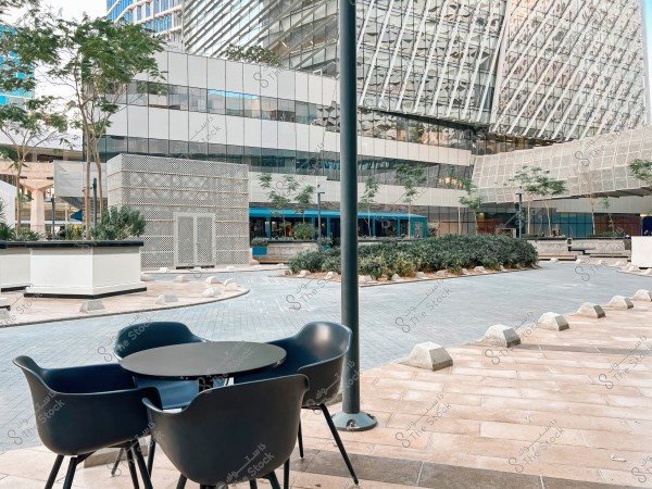 The image shows a modern commercial area with glass buildings featuring contemporary architectural facades. In the foreground, there are four modern-designed chairs and a round table on a paved walkway, surrounded by a few trees and small green spaces. The glass facade of the buildings reflects the sky and intricate geometric designs.