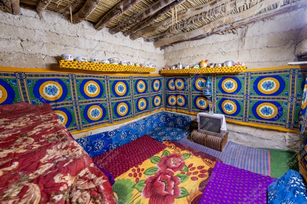 The image shows a traditional room inside a mud brick house. The walls are decorated with colorful fabric featuring blue and yellow floral patterns. There are wooden shelves holding pottery and glassware. The floor is covered with colorful rugs and comfortable cushions, alongside an old television and a lantern. The ceiling is made of wooden logs and palm fronds.