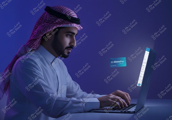 Image of a man sitting and working on a laptop. The man is wearing traditional Saudi attire consisting of a white thobe and a red and white checkered ghutra with a black agal. There is an image of a card labeled \"AlinmaPay\" on the side, suggesting digital transactions or online banking services. The lighting is soft and blue, providing a modern and tech-oriented atmosphere.