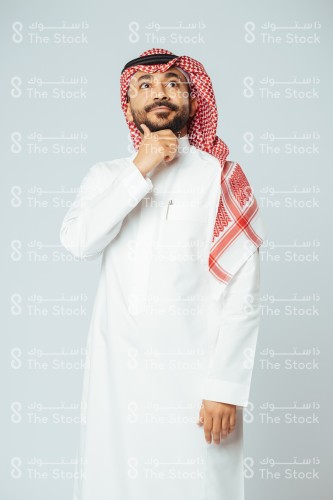 بورتريه رجل سعودي مبتسم يرتدي الزي السعودي التقليدي ويضع يده تحت دقنه بينما ينظر للأعلى وهو يفكر، شاب يتأمل