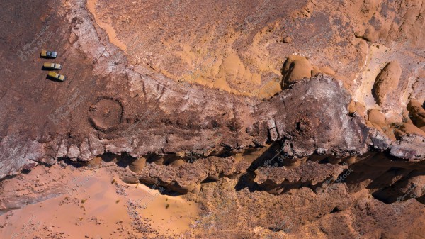 An aerial view of a desert area showcasing undulating, multi-toned brown rocky formations. In the top left corner, three small cars with different colors are parked beside each other. The contrast between the rocky terrain and the desert sands is clearly visible in the image.
