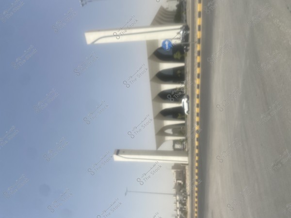 يظهر في الصورة مبنى كبير ذو تصميم معماري حديث مكون من أعمدة ضخمة وسقف مقوس, مع وجود طريق واسع في المقدمة. هناك سيارة بيضاء تقف بالقرب من المبنى ولافتة مرور تشير إلى اليمين, والسماء صافية في الخلفية.