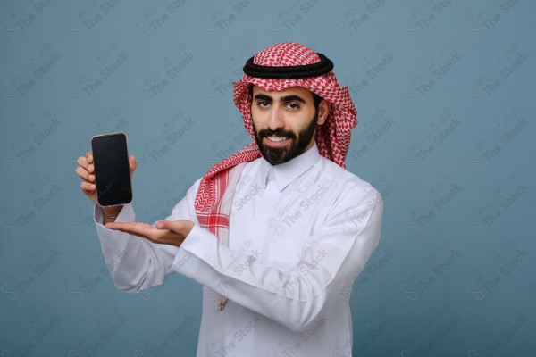 بورتريه شاب سعودي يرتدي الزي السعودي يشير إلى هاتفه ويتفاعل معه، وتظهر على ملامحه الدهشة ، على خلفية سماويه