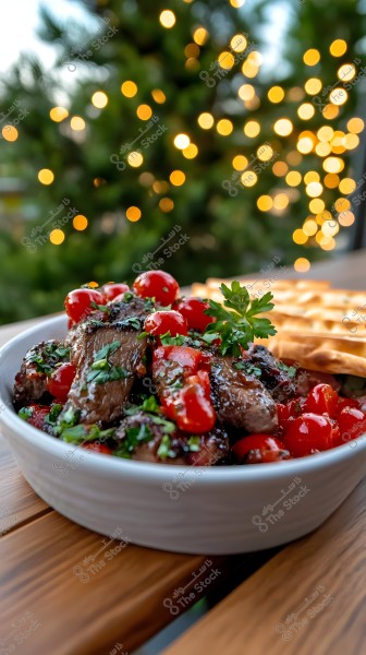 A bowl on a wooden table filled with grilled meat pieces topped with bright cherry tomatoes and fresh parsley leaves, with a beautiful Christmas light background.