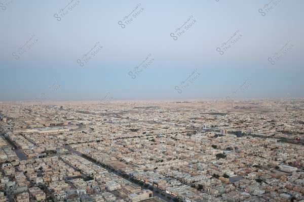 An aerial view of a city showing densely packed buildings stretching to the horizon under a clear blue sky. The streets are straight, dividing neighborhoods filled with low-rise houses and buildings. Some empty spaces can be seen at the horizon, indicating the edge of urban areas.
