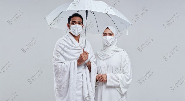 Image of a man and woman dressed in traditional white Ihram clothing. The man is wearing a white wrap around his body and holding a transparent umbrella. The woman is in a white abaya and a white hijab covering her hair, both wearing white medical face masks. The image suggests a religious context such as Hajj or Umrah.
