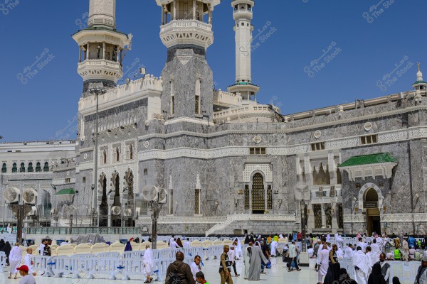 The image shows the exterior courtyard of the Grand Mosque in Mecca, Saudi Arabia, with a large gathering of people wearing traditional white clothing. In the background, parts of the mosque\'s architectural structures can be seen, featuring white marble stones and Islamic decorations. The sky is clear and blue.