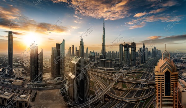 صورة لمدينة دبي في الإمارات العربية المتحدة, تظهر فيها ناطحات السحاب الشاهقة, بما في ذلك برج خليفة في الخلفية. تلتقط الصورة منظرًا بانوراميًا لشبكة الطرق المعقدة في المقدمة, مع شروق الشمس في الأفق مما يضفي وهجًا ذهبيًا على المباني والسماء. السحب الكثيفة تضيف إلى الجمالية الرائعة للمدينة.