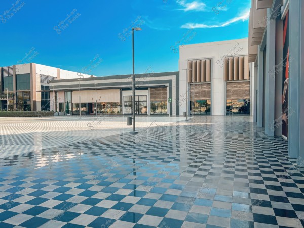 An image of a wide outdoor plaza paved with interlocking tiles in light blue and gray, reflecting light attractively. The surrounding buildings have a modern design, featuring simple architectural details and neutral colors. The sky is blue with light clouds in the background, and there are light poles extending across the plaza.
