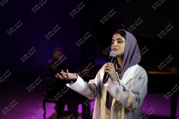 امرأة تغني على المسرح, ترتدي حجابًا أرجوانيًا ومعطفًا أبيض, وتستخدم ميكروفونًا. في الخلفية, يظهر عازف بيانو.