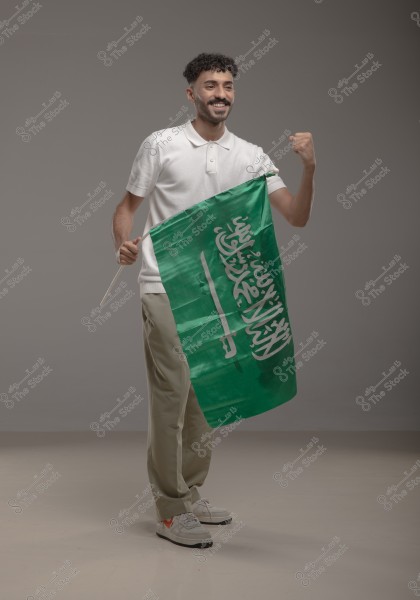 Image of a man standing holding the flag of Saudi Arabia. He is wearing a white polo shirt, beige trousers, and sneakers. The man is smiling and raising his other hand in a fist, expressing pride or happiness. The background is a neutral gray color.