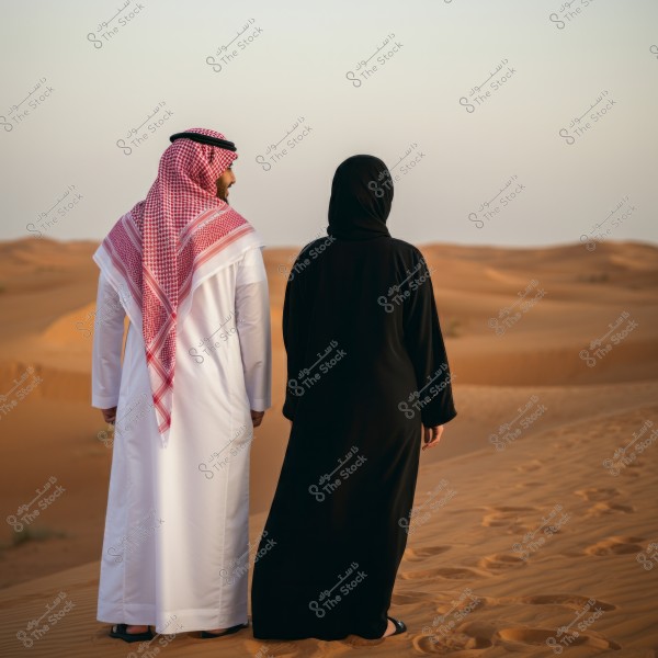 يظهر في الصورة شخصان يقفان في الصحراء. الشخص على اليسار يرتدي ثوبًا أبيض وغترة حمراء وبيضاء مع عقال أسود, بينما الشخص على اليمين يرتدي عباءة سوداء. يمتد خلفهم مشهد صحراوي هادئ يمتاز بالكثبان الرملية الناعمة تحت السماء الصافية.