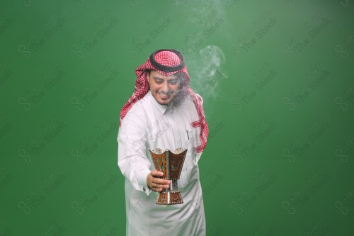 رجل سعودي يرتدي الزي التقليدي على خلفية خضراء ، التطيب بالبخور والعود ، استخدام خشب العود في المناسبات والأعياد.