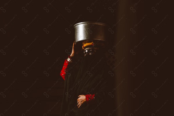 A picture of a woman wearing the traditional dress that represents the Saudi culture, carrying a pot over her head, the traditional dress.