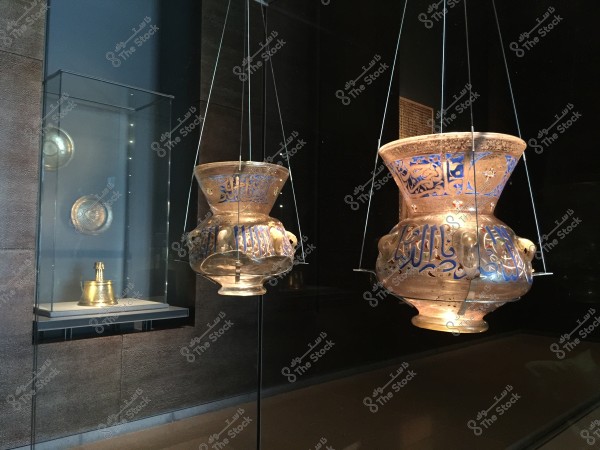 An image of two blown-glass lamps with intricate Islamic designs and Arabic inscriptions, displayed in a museum setting. The lamps hang from the ceiling by thin wires and exhibit a golden hue with blue details. In the background, there is a glass display case with round golden metal artifacts.