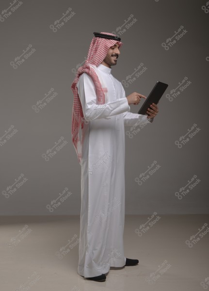 Image of a man wearing a white thobe and a red and white headscarf (ghutra) with a black agal. The traditional clothing suggests he is from Saudi Arabia. The man is using a tablet with one hand and smiling in a relaxed atmosphere in a simple studio with neutral colors.