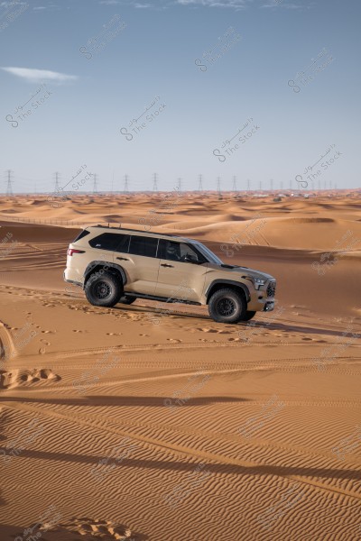 A white SUV driving across sand dunes in the desert during the daytime. The background displays a vast scene of sand with power lines stretching along the horizon. The sky is clear blue.