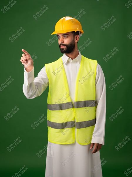 رجل يرتدي خوذة بناء صفراء وسترة عاكسة يقف في وضع يشير بيده اليمنى, مع خلفية خضراء.
-
