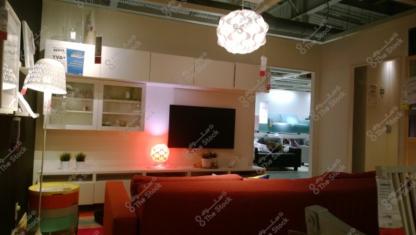 A modern living room with a red sofa facing a wall-mounted TV. Next to the TV, a glowing lamp emits a red hue, and a white table holds small potted plants. The back wall features white cabinets and modern pendant lights. In the background, a furniture store display section shows sofas and other furniture items.