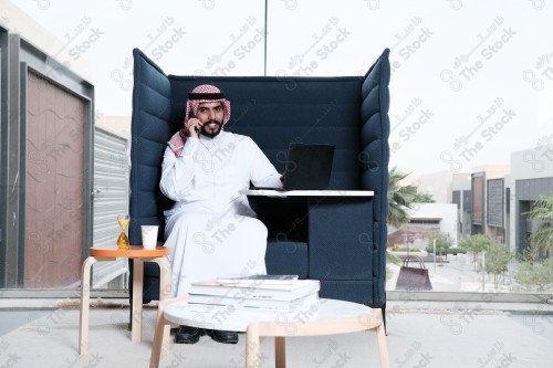 رجل سعودي يرتدي الزي السعودي التقليدي يقوم بإجراء مكالمة هاتفية بينما يدون ملاحظاته على إحدى صفحات المفكرة ويظهر الجهاز المحمول على سطح الطاولة أثناء النهار