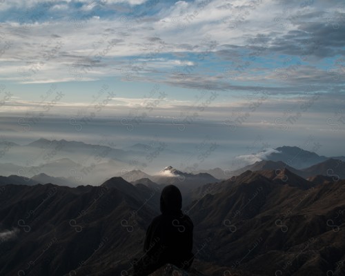 A shot of a man standing on the edge of the mountain at sunrise from the heights of the Asir Mountains, clouds embrace the top of the mountains, a chain of rocky mountains in the Asir region.