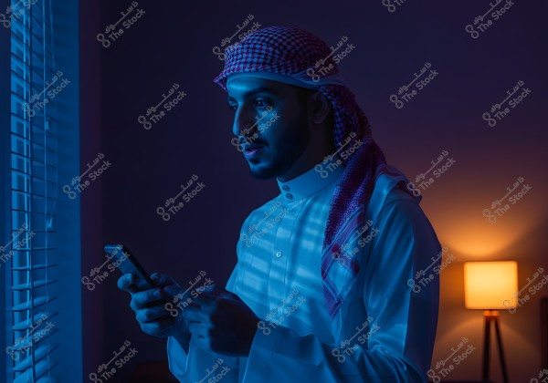 A man wearing traditional Gulf attire stands by a window holding a mobile phone. The room is dimly lit with some light coming from a lamp in the background, creating a calm atmosphere. The man is wearing a white and red checked headscarf and a white thobe.
