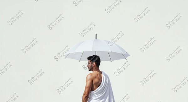 Image of a shirtless man standing at a side angle, holding a large white umbrella. He is draped with a white cloth covering part of his back and shoulder. The background is white and clean, creating a minimalist and pure look.