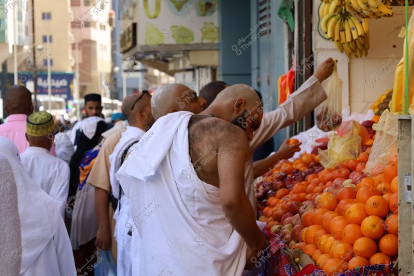The image shows several people wearing white Ihram garments, standing in front of a fruit vendor in an open market. The vendor displays a variety of fruits such as oranges, apples, and bananas. In the background, there are multi-story buildings and crowded streets, suggesting the scene might be in a bustling city, possibly Mecca in Saudi Arabia, where people wear Ihram clothing.