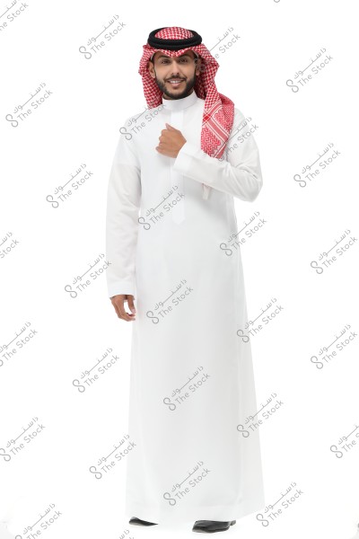 A portrait of a smiling man standing and wearing traditional Saudi Arabian white thobe along with a red and white checkered shemagh and a black igal. The image is taken against a white background.