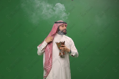 رجل سعودي يقف على خلفية خضراء يرتدي الثوب و الشماغ، يقوم بعمل حركات بيديه ، يبخر نفسه بالطيب و العود، احتفالات العيد