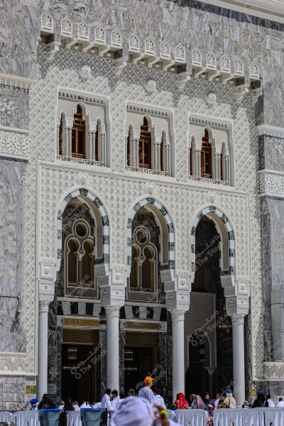 An image of an ornate and luxurious architectural façade featuring Islamic geometric designs including arches, columns, and windows. The top of the façade is adorned with diamond shapes and intricate carvings, while the windows are framed by small columns. Below, a group of people are seen wearing traditional Ihram clothing in the holy mosque area in Mecca.