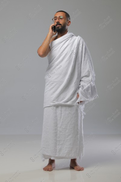 صورة لرجل يرتدي ملابس الإحرام البيضاء, وهو يتحدث على الهاتف المحمول. يظهر الرجل في وضعية وقوف مع نظرة مركزة إلى الأمام, ويرتدي نظارات. تظهر أرضية وجدار خلفية رمادي فاتح.