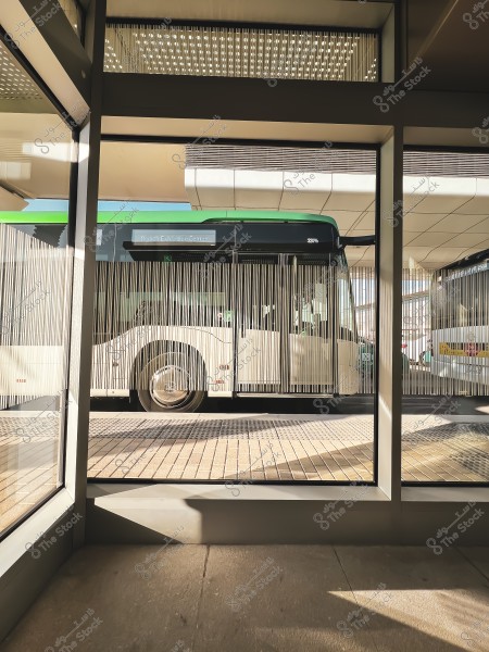 Image showing a white bus with a green roof passing by a modern bus station. The station\'s interior features a contemporary design with large glass windows allowing natural light to enter. The digital sign on the bus indicates it\'s heading to \"Riyadh Exhibition Center.\"