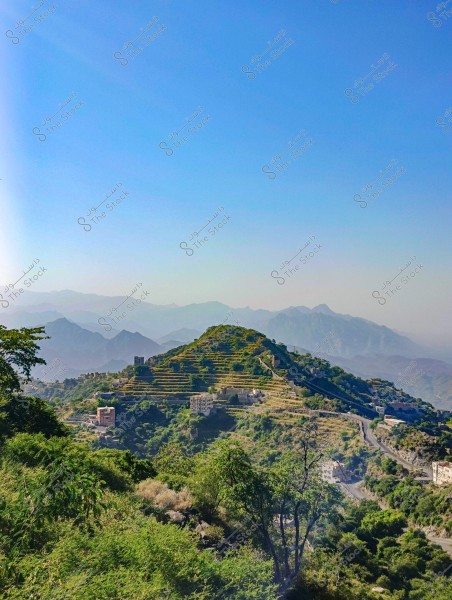 A landscape view of a mountainous area featuring green agricultural terraces and scattered houses among the hills. A range of mountains can be seen in the distance, with a clear blue sky stretching across the entire scene.