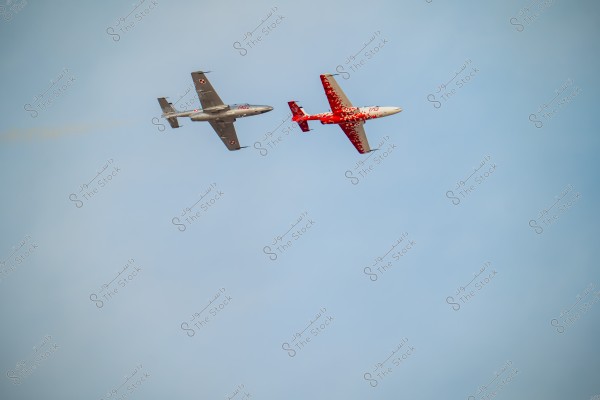 Two jet aircraft flying close to each other in a clear blue sky. The aircraft on the left is silver, while the one on the right is red with a white design.