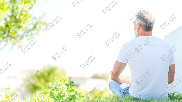 رجل جالس على العشب في حديقة طبيعية, يرتدي قميصًا أبيض وسروال جينز أزرق فاتح. يظهر ظهره فقط, مع توزيع الشعر الأبيض والرمادي. في الخلفية, تظهر الأشجار الخضراء والسماء الصافية.