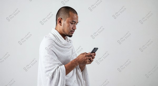 An image of a man wearing a white wrap, seemingly in a state of Ihram, holding a mobile phone and looking at it, against a simple white background.