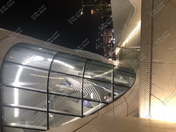 مبنى حديث ذو تصميم مستقبلي مائل قليلاً في الليل, يتميز بنافذة زجاجية كبيرة ومتعرجة تتميز بإضاءة داخلية زاهية. في الخلفية, يمكن رؤية ممر مضاء بالأضواء وخلفه مشاهد من المدينة في الليل مع أضواء السيارات والمباني. يظهر التصميم باللون الأبيض والخطوط النظيفة.