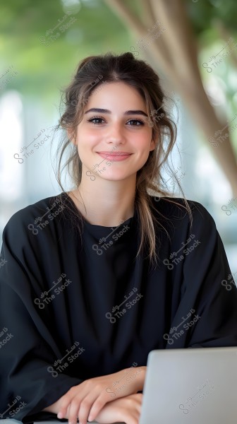 صورة لامرأة ترتدي ملابس سوداء, جالسة وأمامها جهاز كمبيوتر محمول. تبدو مبتسمة وذات مظهر هادئ. الخلفية غير واضحة, مما يركز الانتباه على المرأة. البيئة المحيطة تبدو وكأنها في مكان مغلق مع بعض الديكورات الطبيعية الخضراء.