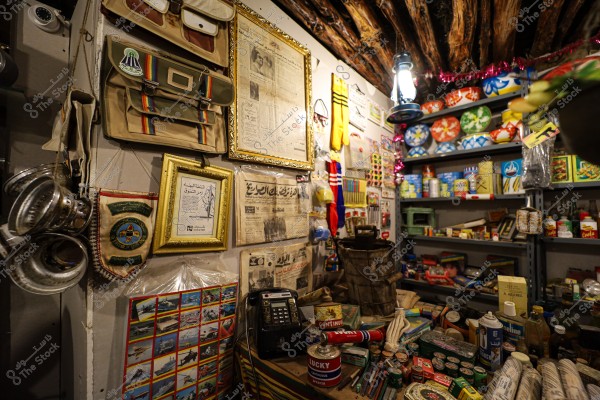 An image of an old shop displaying a collection of vintage tools and antiques. The image shows various cans and old products on metal shelves, including paint tins, oils, and some food products. The walls are adorned with old newspapers, framed historical pictures, and hanging vintage bags, along with colorful decorations. A traditional lantern illuminates the space, and there is an old telephone and wooden tables displaying assorted tools.
