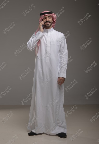 A portrait of a man standing and smiling while talking on a mobile phone. He is wearing a white thobe and a red and white checkered headscarf with a black agal, indicative of traditional attire from the Gulf region, possibly Saudi Arabia. The setting is indoors with a neutral background.