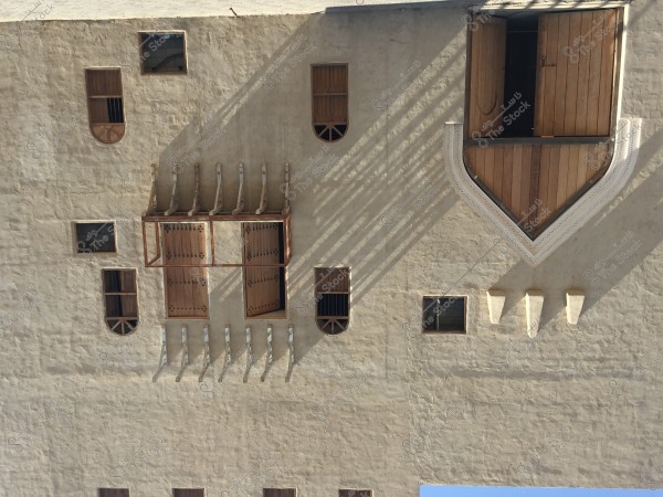 An image of a traditional building wall with plaster facades and large wooden doors. The wall features several rectangular wooden windows and round windows at the edges with intricate decorations. The design showcases traditional architectural elements and might be from a region in the Arabian Gulf, such as Saudi Arabia.