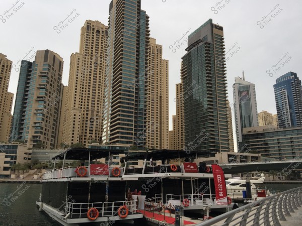 The image shows a marina area with multiple skyscrapers surrounding it. The buildings are tall with glass facades and lighting. In the foreground, there is a luxury yacht labeled \"Xclusive Yachts\" docked at the marina. The sky is overcast, giving a calm atmosphere.
