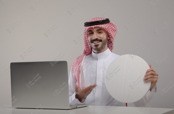 Image of a man wearing traditional Saudi attire, including a white thobe and red and white checkered headscarf, sitting in front of a laptop and holding a round, white board. The man is smiling in a simple indoor setting.