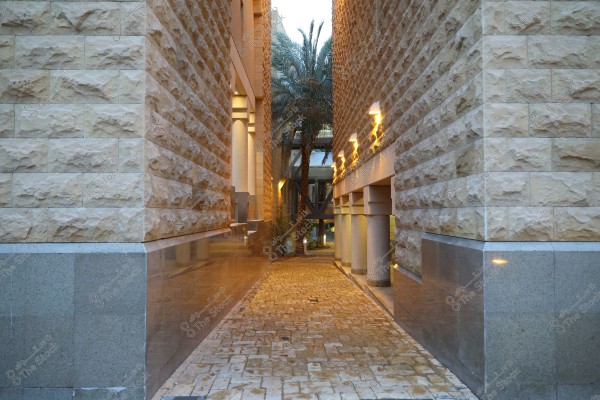 A narrow passageway between two buildings with beige stone walls featuring a modern architectural design. A palm tree stands in the middle of the passage, and lights on the walls create a warm ambiance. The floor is paved with light brown stones.