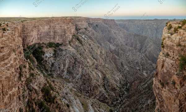 A panoramic image of a deep rocky valley in a mountainous region, with steep rock walls extending across the scene. Sparse green vegetation is visible along the rocky slopes, and the sky is clear and calm on the horizon. The geographical depth of the scene highlights the rugged beauty of the landscape.