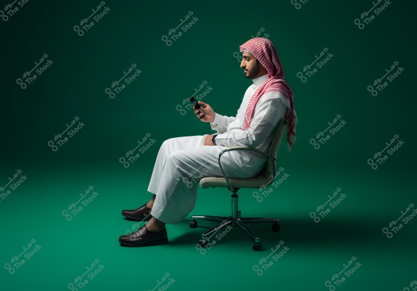 صورة لرجل يرتدي ثوباً أبيض وغترة حمراء جالس على كرسي دوار, يمسك بهاتف محمول في يده. خلفية الصورة خضراء, مما يبرز ملامح الرجل وملبسه التقليدي, الذي يشير إلى أن موطنه قد يكون في المملكة العربية السعودية.